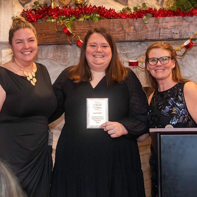Angelia Miller centered holding an award with a female presenter standing on each side 